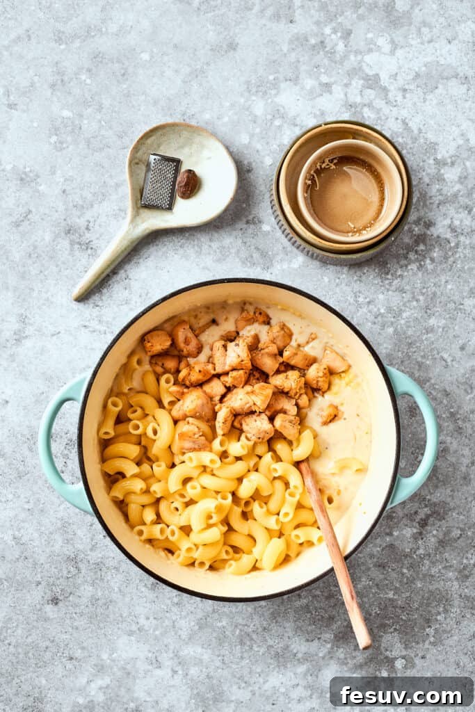 Cooked chicken pieces are being stirred into a large pot of creamy macaroni and cheese, ensuring every bite is hearty.