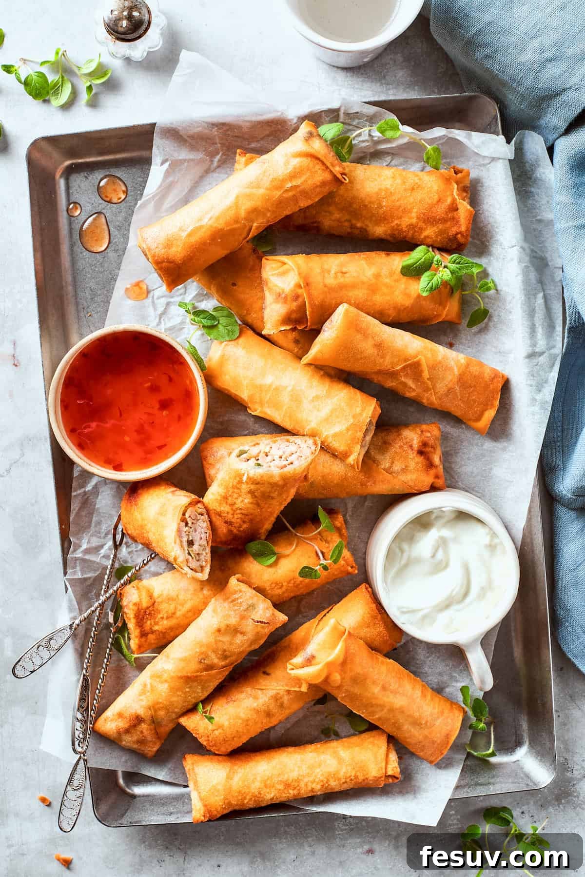 A serving tray filled with golden-brown crispy chicken egg rolls, accompanied by small bowls of vibrant red sweet chili sauce and creamy white dipping sauce.