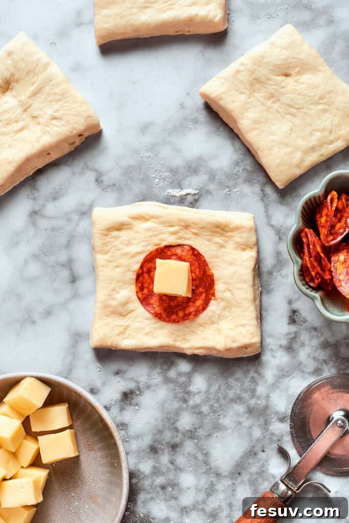This easy Pizza Bites recipe stuffs spicy pepperoni and gooey mozzarella cheese into little pieces of pizza dough brushed with herb butter. A single slice of pepperoni and a small cube of cheese are carefully placed in the center of a pizza dough square, ready for folding.