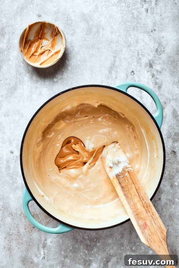 No-bake Peanut Butter Rice Crispy Treats are swirled with creamy peanut butter and capped with a rich layer of melted milk chocolate. Creamy peanut butter is being stirred into the smooth, melted marshmallow mixture in a saucepan.