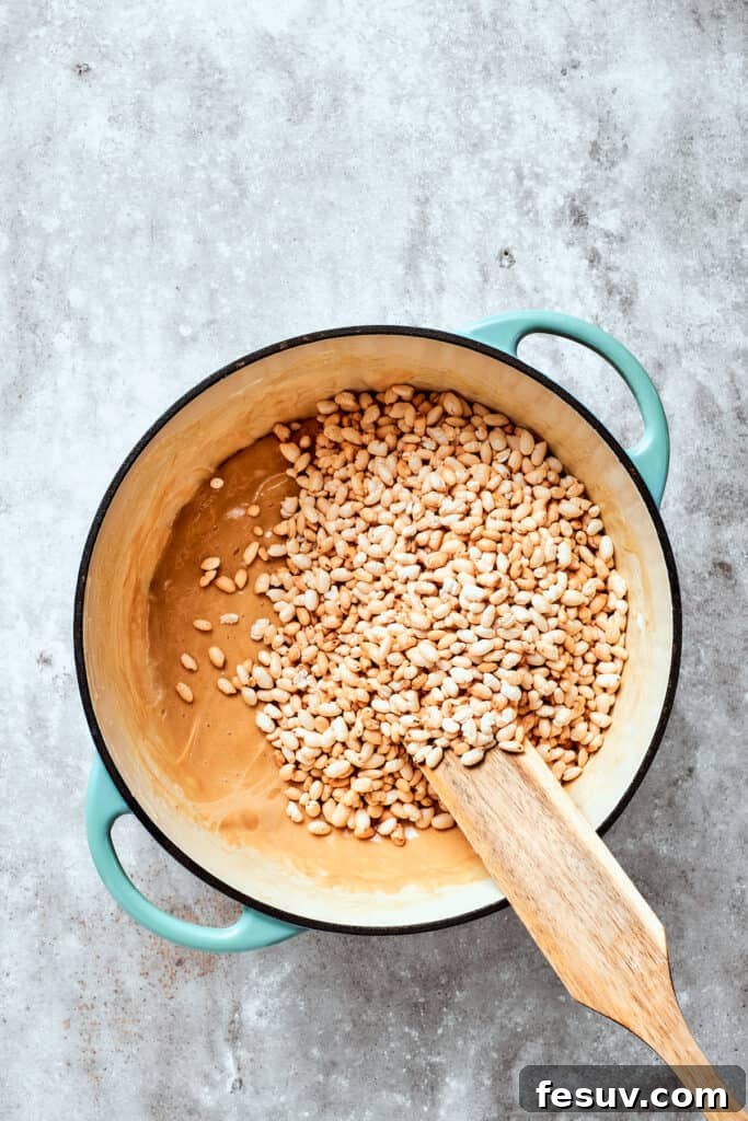 No-bake Peanut Butter Rice Crispy Treats are swirled with creamy peanut butter and capped with a rich layer of melted milk chocolate. Crispy rice cereal is being gently stirred into the peanut butter and marshmallow mixture, ensuring every piece is coated.