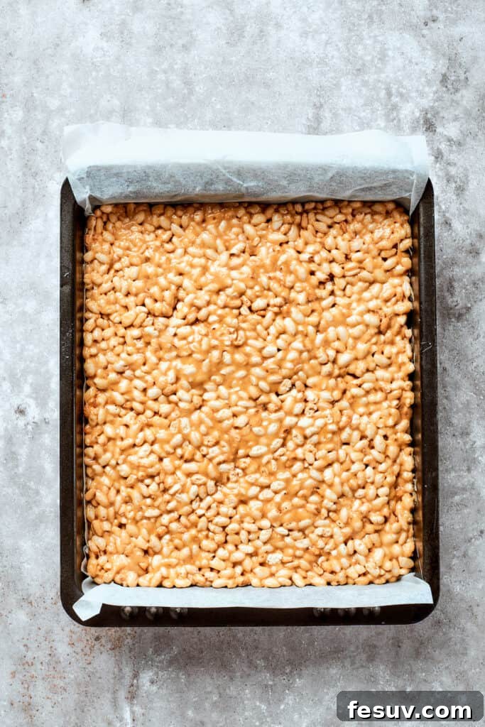 No-bake Peanut Butter Rice Crispy Treats are swirled with creamy peanut butter and capped with a rich layer of melted milk chocolate. The peanut butter rice crispy treat mixture is being pressed firmly and evenly into a parchment-lined square baking dish.