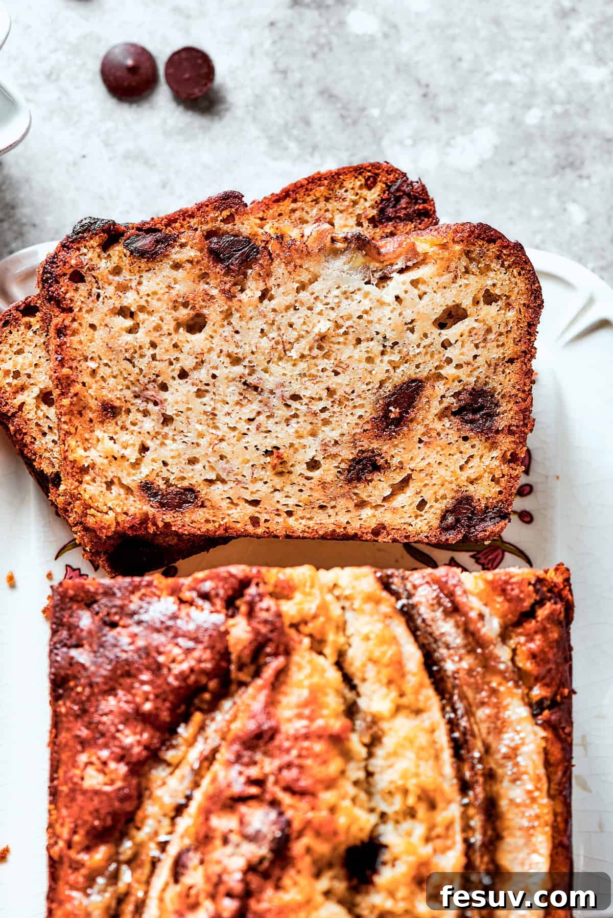 An overhead view of a beautifully baked sour cream banana bread, with two generous slices cut out, revealing its moist crumb, decorative banana top, and scattered chocolate chips.