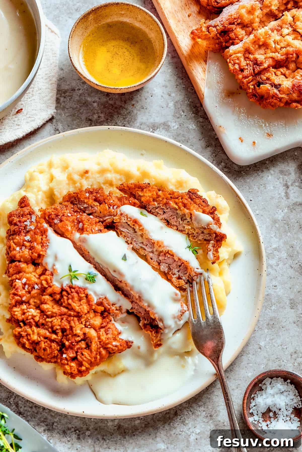 A mouthwatering serving of chicken fried steak with mashed potatoes. Sliced chicken fried steak served over mashed potatoes with a fork, drenched in gravy.