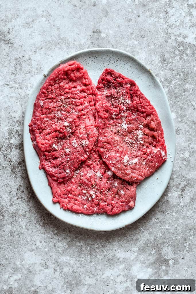 Seasoning the cube steaks evenly for maximum flavor. Three pieces of cube steak seasoned on a plate.