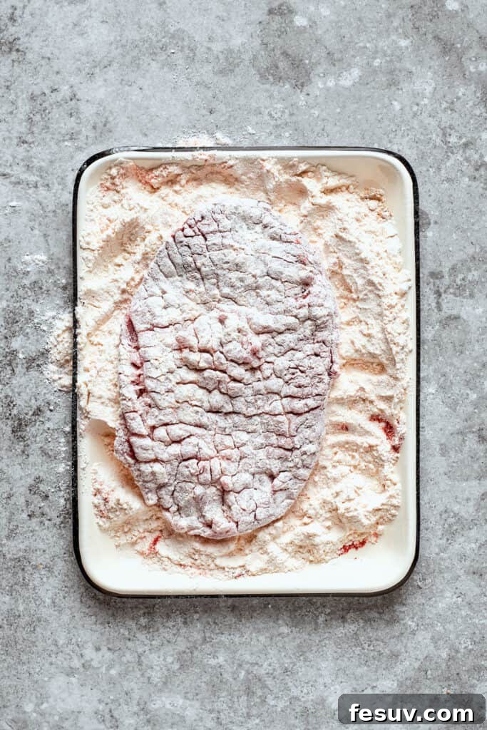 First dredge: Coating the steak thoroughly in seasoned flour. A cube steak being coated in flour from both sides.