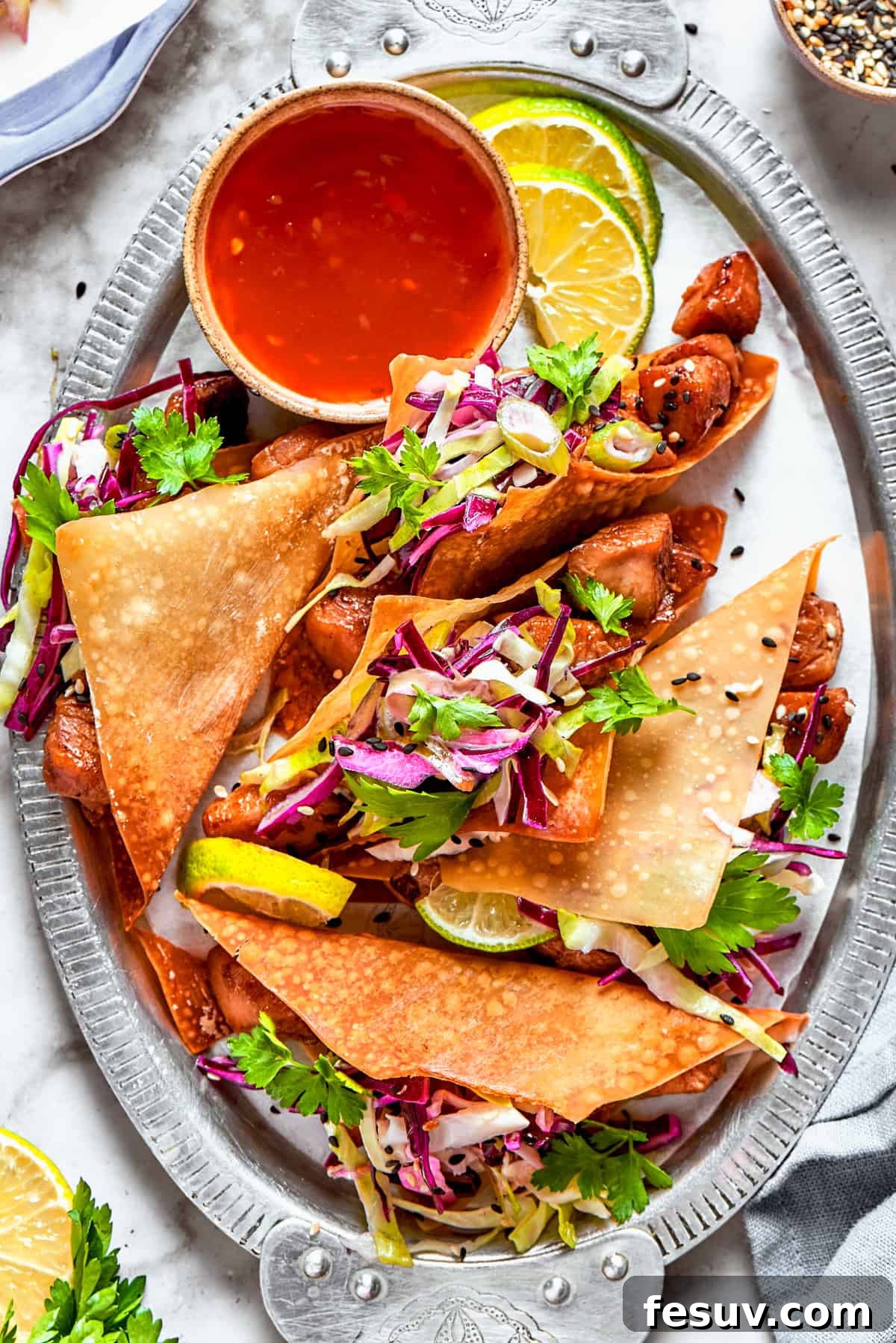 These Chicken Wonton Tacos feature bites of garlicky honey chicken stuffed into crisp wonton shells along with tangy fresh coleslaw. Chicken wonton tacos arranged on an oval platter with a bowl of sauce set right next to the tacos.