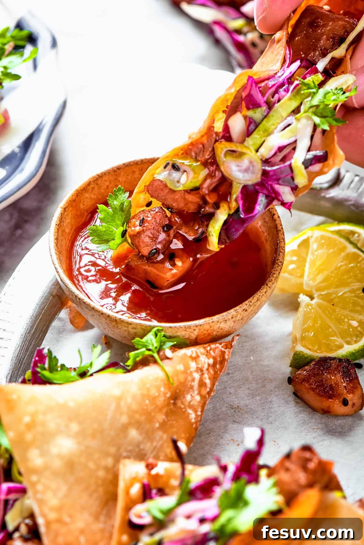 These Chicken Wonton Tacos feature bites of garlicky honey chicken stuffed into crisp wonton shells along with tangy fresh coleslaw. Dipping a taco into a bowl of red sauce.
