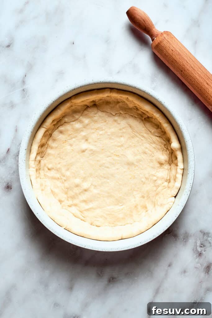Press the deep dish pizza dough firmly into the prepared pan. The rolled-out deep dish pizza dough is carefully pressed into a deep-sided baking pan, forming the base and sides of the pizza, ready for toppings.