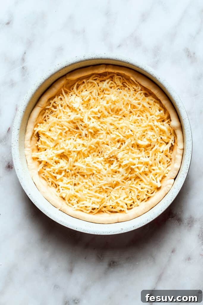 Add your shredded cheeses as the first layer on the deep dish crust. A generous layer of shredded mozzarella and Edam cheese covers the deep dish pizza dough, forming the foundational cheese layer before the sauce is added.