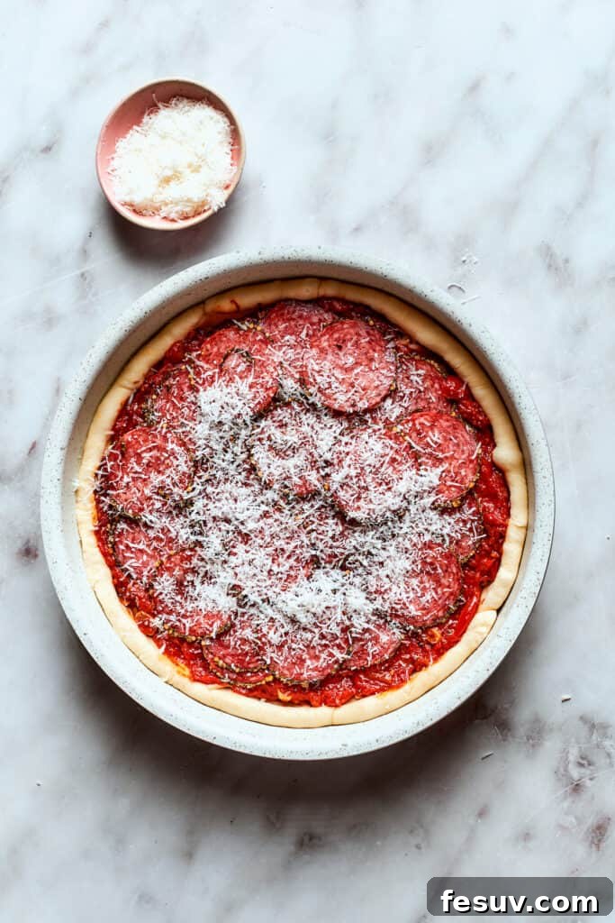 Sprinkle Parmesan cheese and arrange pepperoni on top of the sauce. Grated Parmesan cheese is generously sprinkled over the deep dish pizza sauce, adding a final savory touch before the pepperoni and baking.