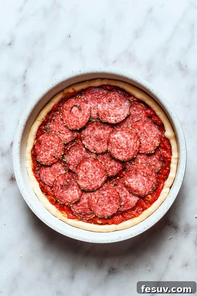 The assembled deep dish pizza is ready to be baked to golden perfection. A deep dish pizza, fully assembled with layers of crust, cheese, sauce, pepperoni, and Parmesan, is perfectly ready to be baked to golden perfection.