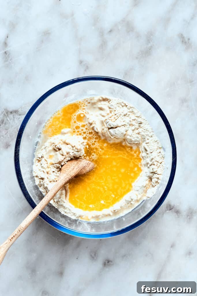 Incorporate wet ingredients into the deep dish pizza dough mixture. Warm water and melted butter are being stirred into the dry ingredients for the deep dish pizza dough, starting to form a cohesive mixture.