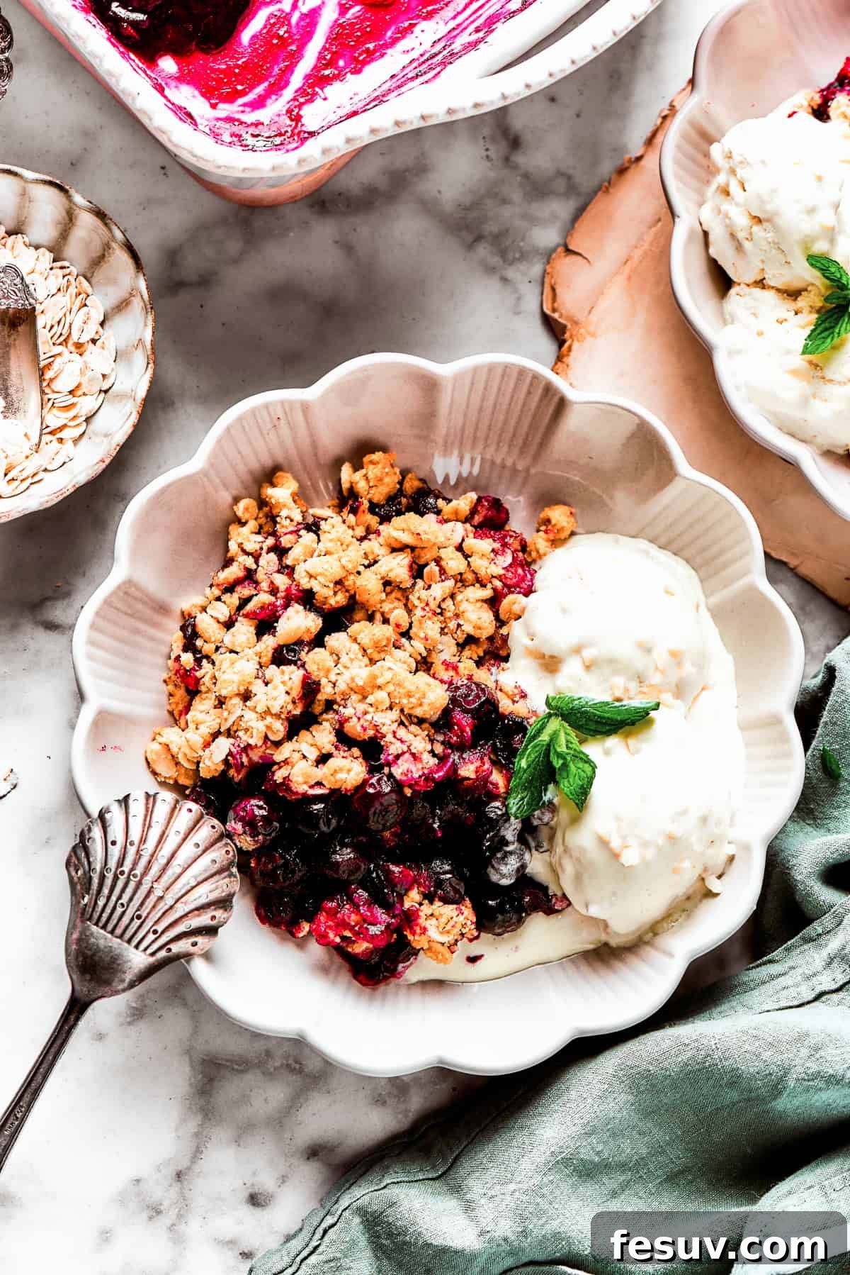 Blueberry crisp served with ice cream and mint garnish A serving of warm blueberry crisp in a bowl, topped with two scoops of vanilla ice cream, garnished with a fresh mint leaf, and ready to be enjoyed.