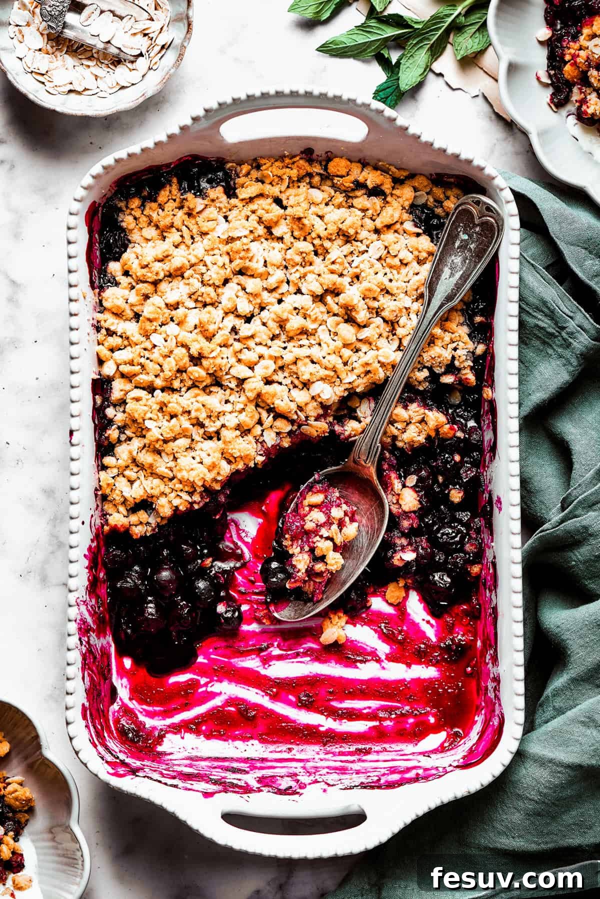 Freshly baked blueberry crisp with a scoop removed An overhead shot of a baked blueberry crisp in a ceramic baking dish, with a section scooped out, revealing the warm, jammy blueberry filling underneath the golden, crunchy topping.