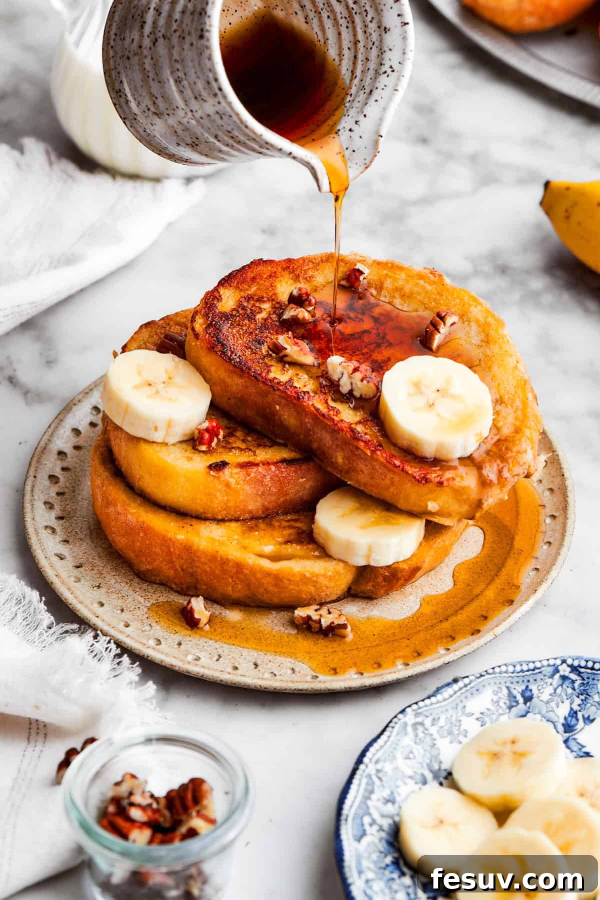 Golden Banana French Toast generously drizzled with maple syrup. A gleaming pitcher elegantly pours rich, amber maple syrup over a towering stack of freshly prepared banana French toast, creating a tempting cascade.