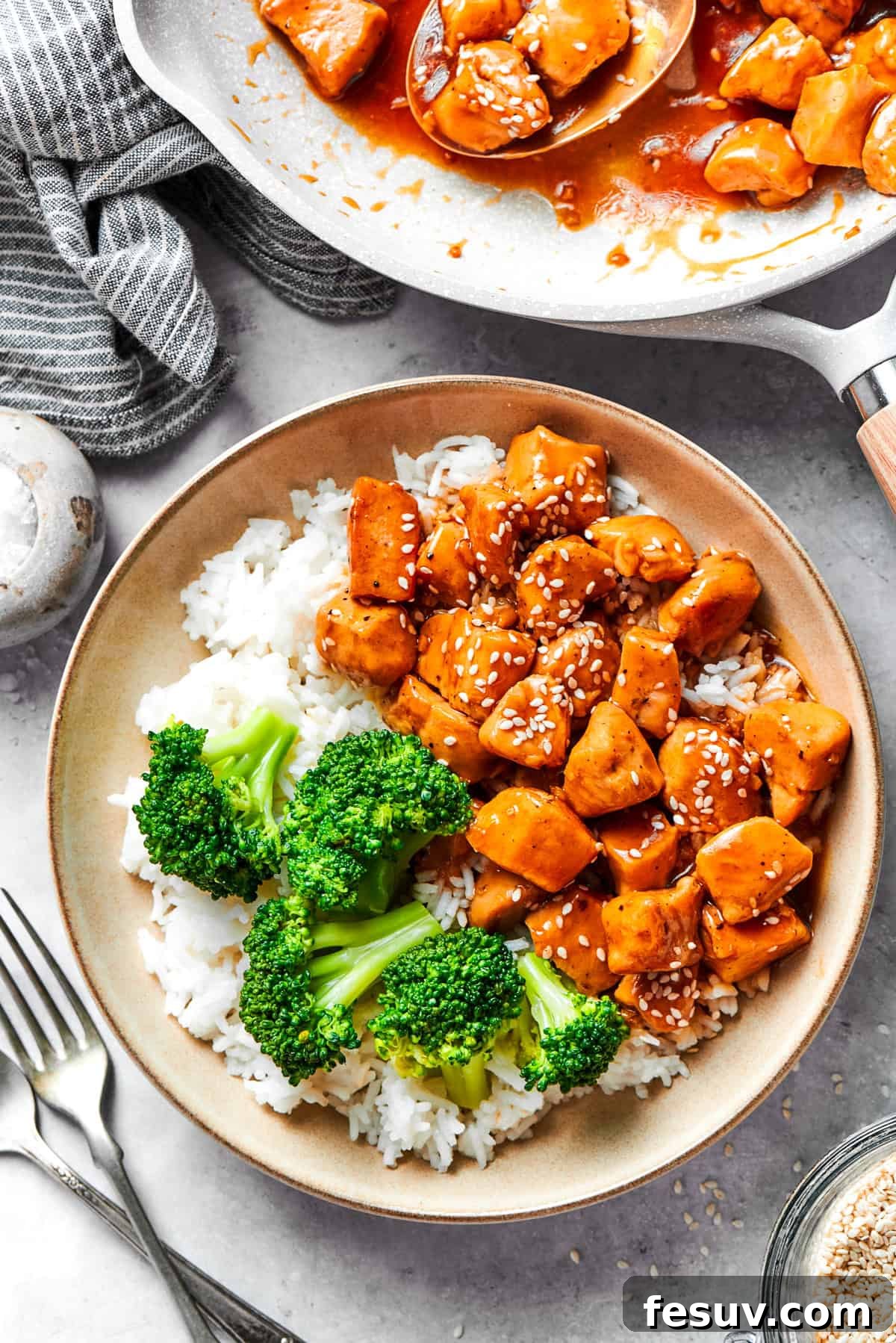 A bowl of rice is topped with teriyaki chicken and broccoli with more teriyaki chicken in the background.