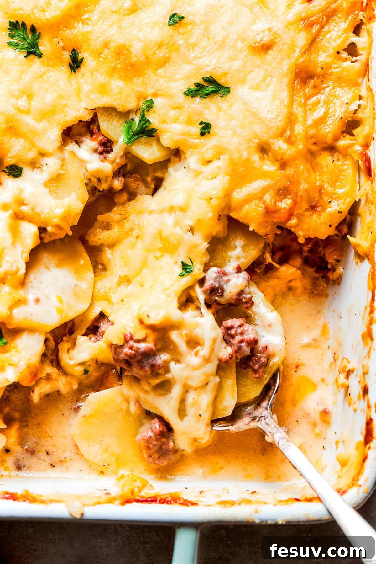 A close-up view of Hamburger Potato Casserole, highlighting its creamy texture with savory ground beef, tender potatoes, and gooey melted cheese. A serving spoon expertly mixes through a rich, bubbly Hamburger Potato Casserole, showcasing the layers of perfectly cooked ground beef and tender sliced potatoes beneath a golden, melted cheese topping.