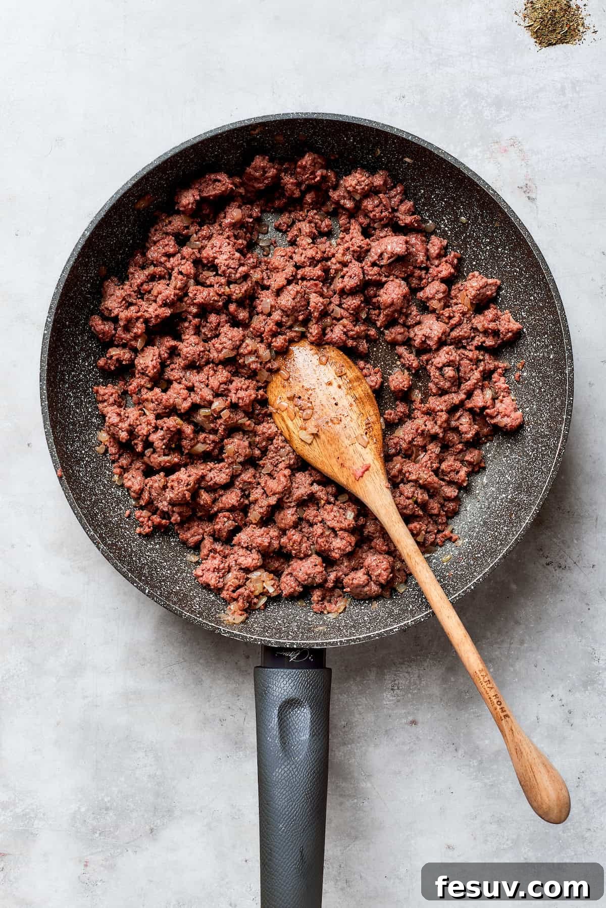Browned ground beef in a skillet, ready for the next step in the casserole. The ground beef is actively browning in the skillet alongside chopped onions, breaking apart into fine crumbles and releasing a rich aroma.