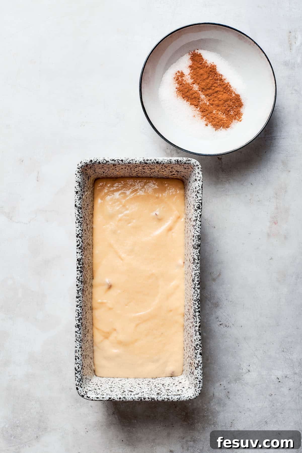 Amish friendship bread batter in a loaf pan, with a small bowl of cinnamon sugar topping beside it.
