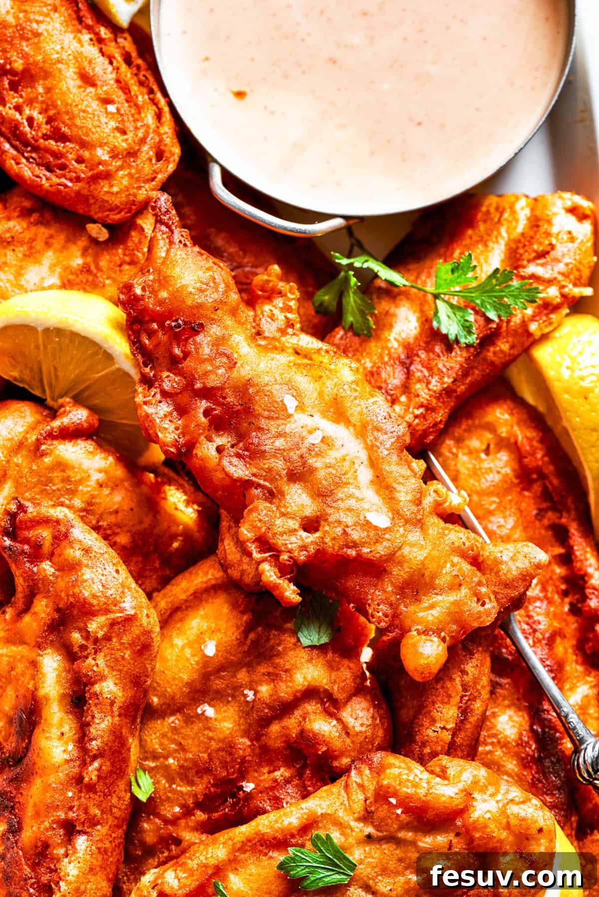 A close-up photo of beautifully golden and crispy beer battered chicken pieces arranged artfully on a plate with a small bowl of spicy dipping sauce.