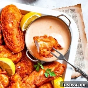 A piece of beer battered chicken is dipped into spicy sauce with more chicken pieces around it.