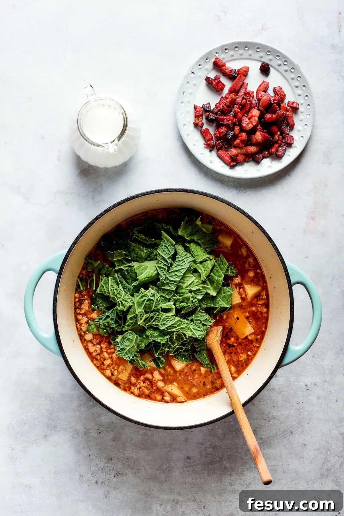 My Zuppa Toscana recipe features Italian sausage, bacon, potatoes, and kale in a savory broth. It’s an easy copycat version of the Olive Garden classic! Freshly chopped green kale leaves carefully added to the top of a simmering soup pot, ready to be stirred in.