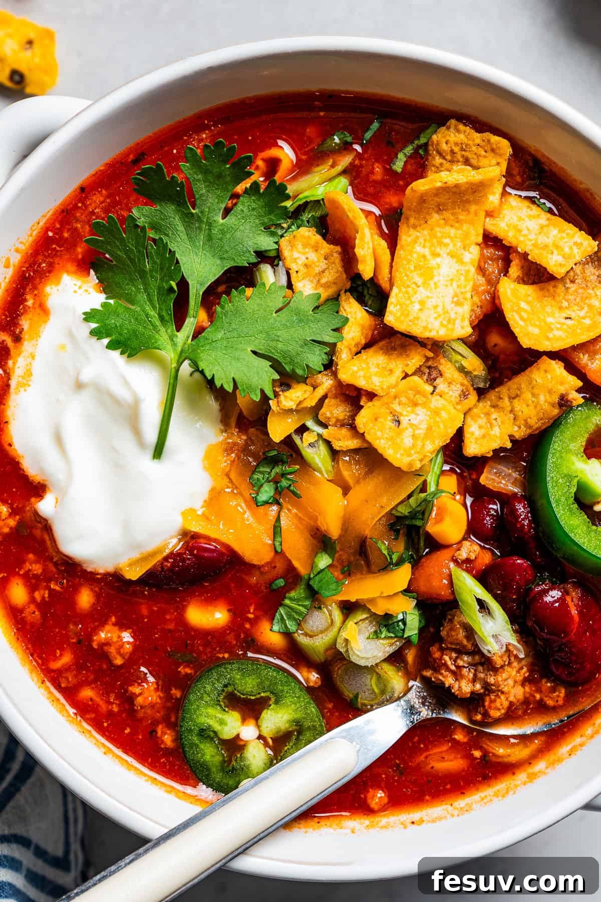 The BEST turkey chili recipe with juicy ground turkey, veggies, beans, and the perfect blend of spices. Great for weeknights and game days! Overhead close up view of a bowl of turkey chili garnished with generous dollops of sour cream, vibrant green jalapeños, and crunchy corn chips.