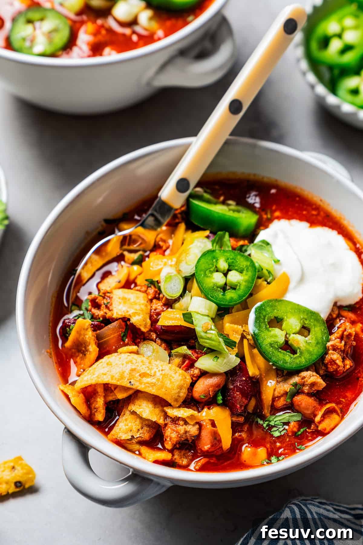 The BEST turkey chili recipe with juicy ground turkey, veggies, beans, and the perfect blend of spices. Great for weeknights and game days! A beautifully garnished bowl of turkey chili with sour cream, jalapeños, and corn chips, with a second, slightly blurred bowl in the background, creating a cozy scene.
