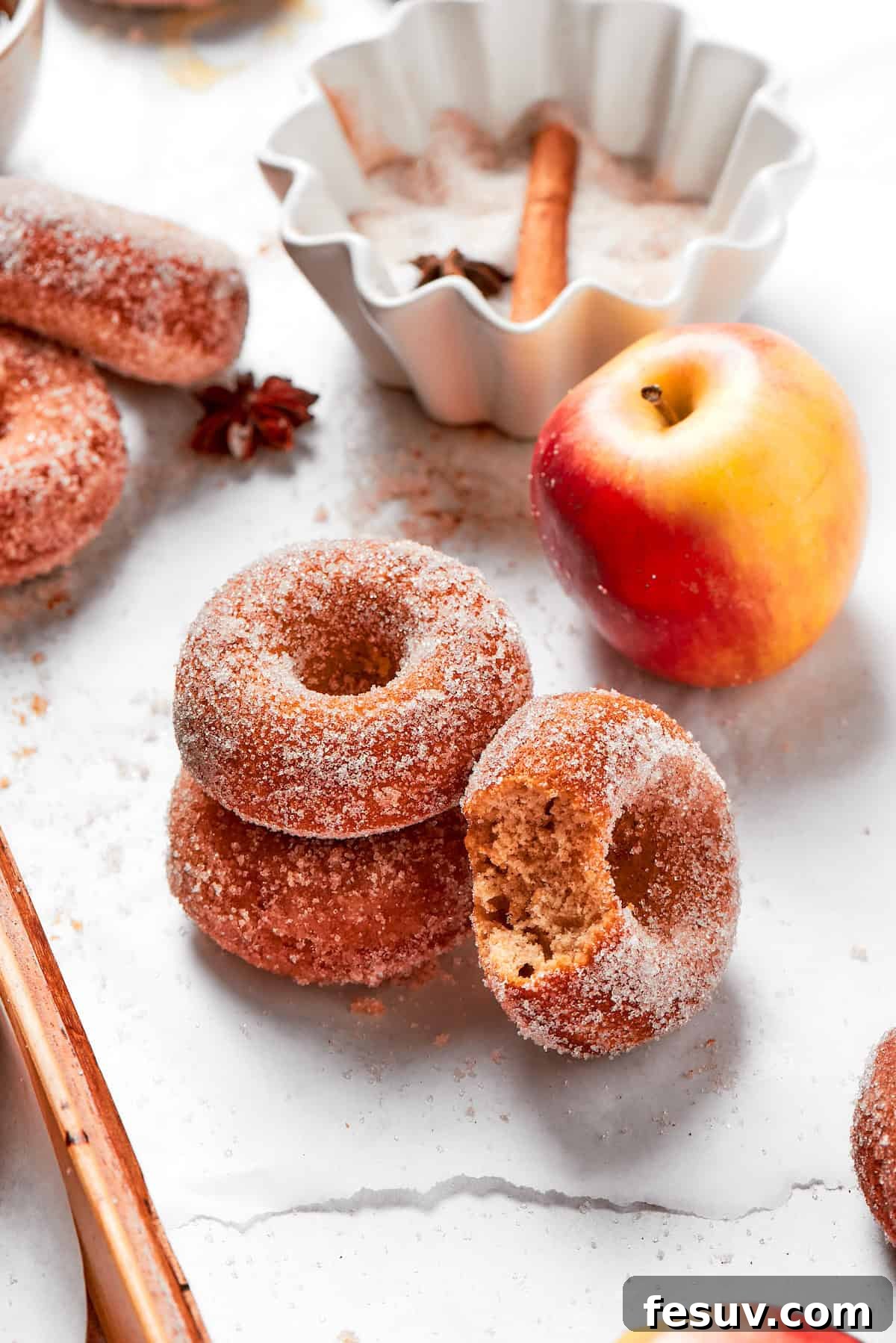 A small stack of perfectly baked apple cider donuts, glistening with spiced sugar, beside a fresh, crisp apple, evoking the cozy flavors of autumn.