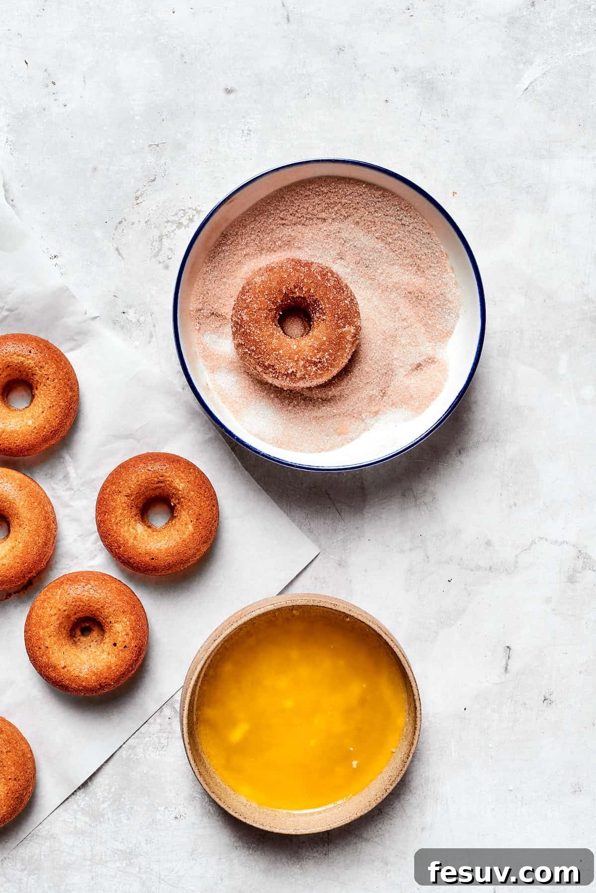 A freshly baked apple cider donut is being dipped into a bowl of sparkling cinnamon sugar after being brushed with melted butter, ensuring a generous coating.