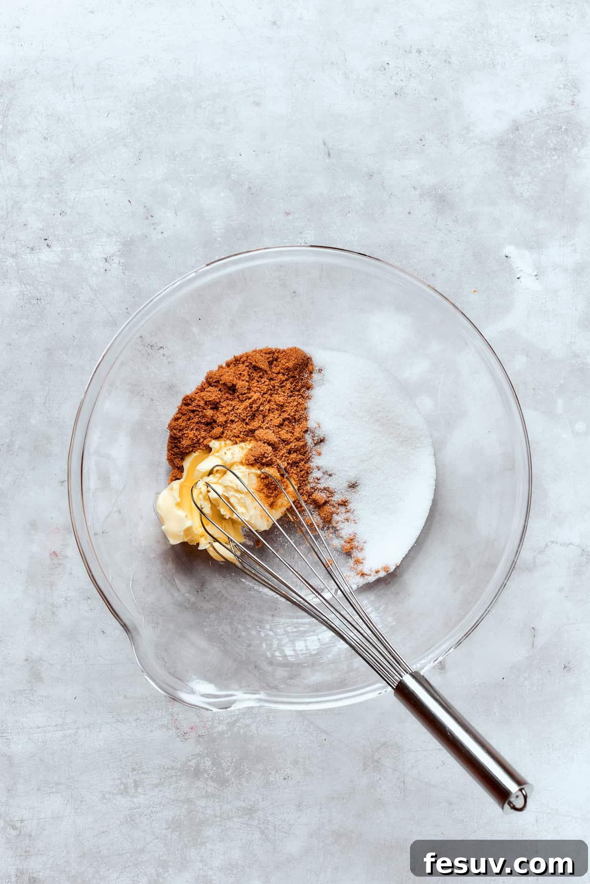 A whisk vigorously mixes softened butter with granulated and brown sugar in a large mixing bowl, creating a light and fluffy mixture.