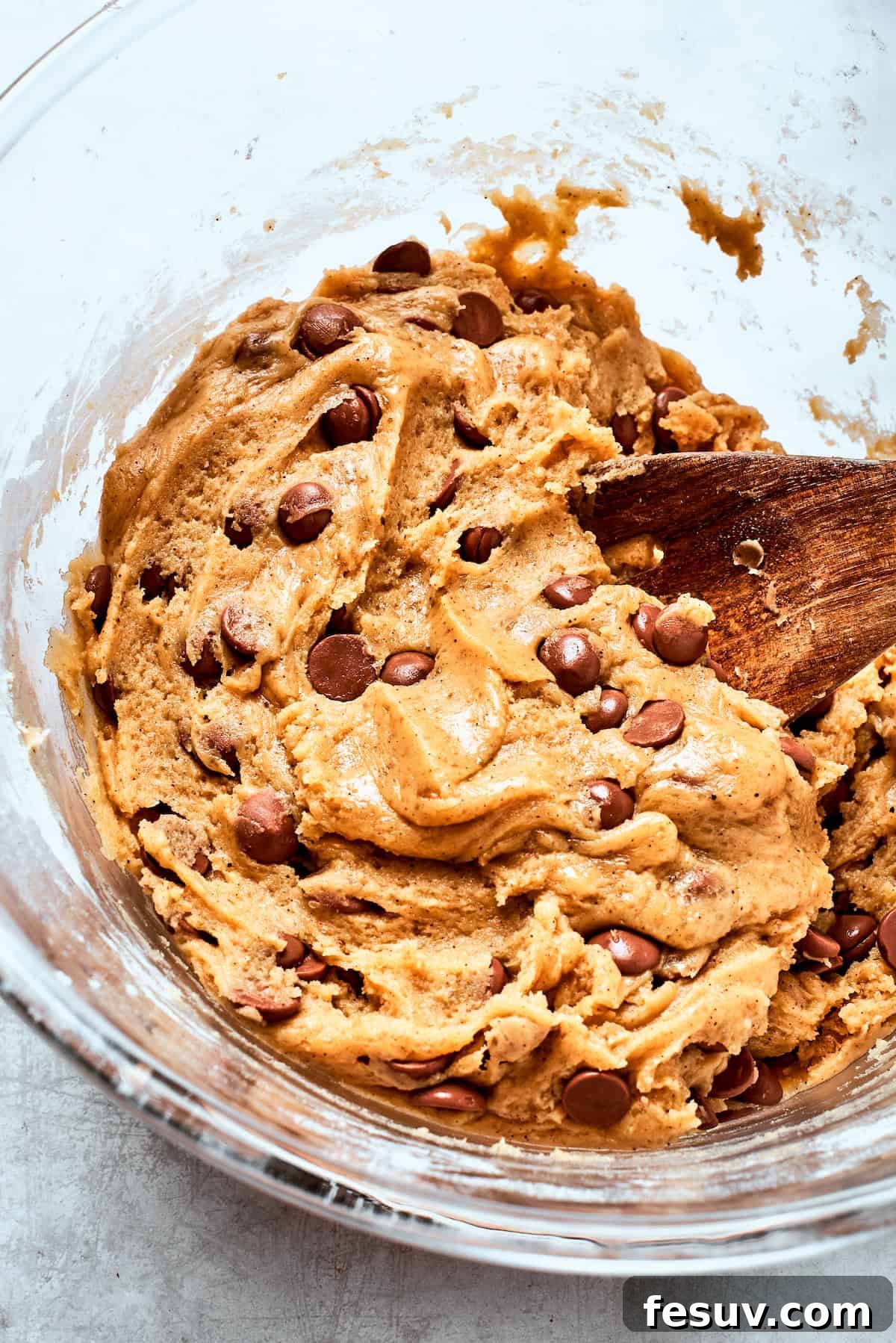 Semi-sweet chocolate chips being folded into a bowl of rich brown butter cookie dough with a wooden spoon.