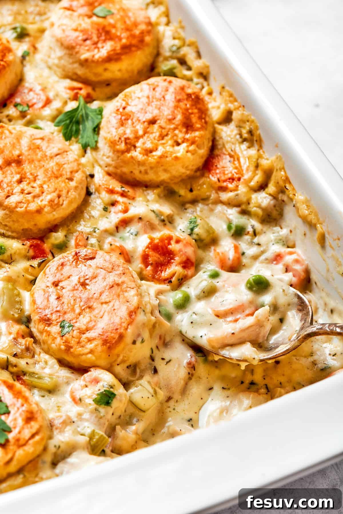 Chicken pot pie casserole is everything you love about the classic comfort food, baked under fluffy buttermilk biscuits instead of pie crust! A golden-brown chicken pot pie casserole in a white baking dish, fresh from the oven, with a spoon taking out a portion.