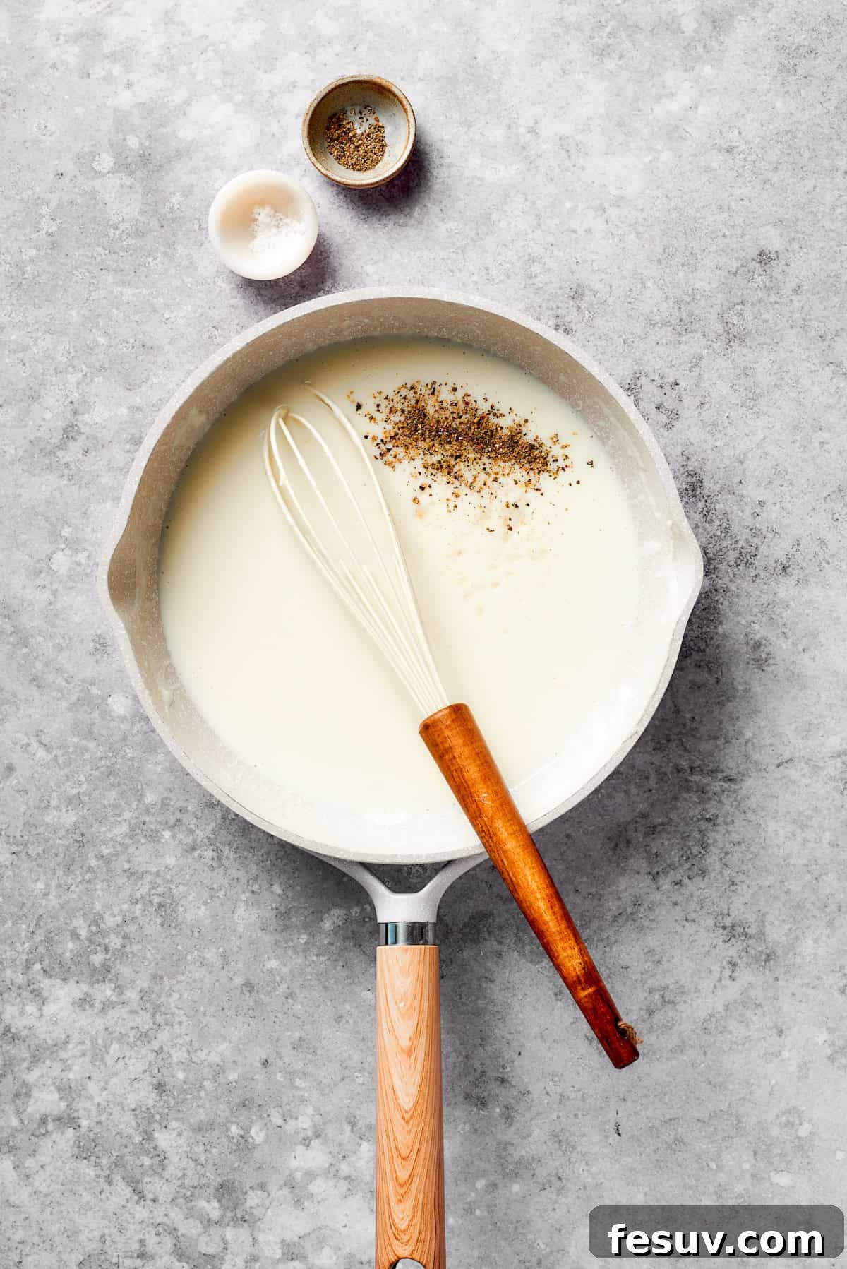 Milk is gradually added to the roux in a saucepan, whisked continuously, and seasoned with pepper to create a smooth béchamel sauce.