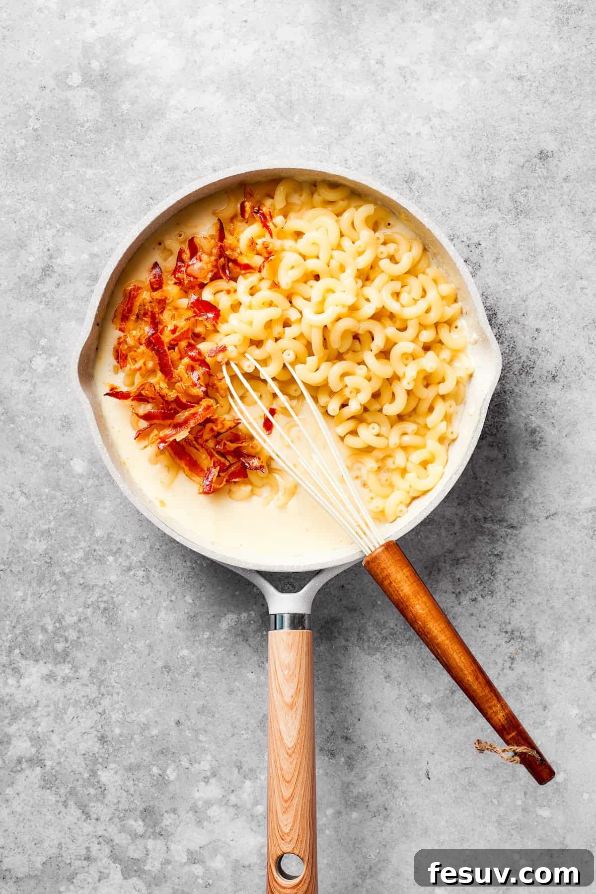 Cooked macaroni and crispy bacon are being stirred into the creamy cheese sauce in a large saucepan.