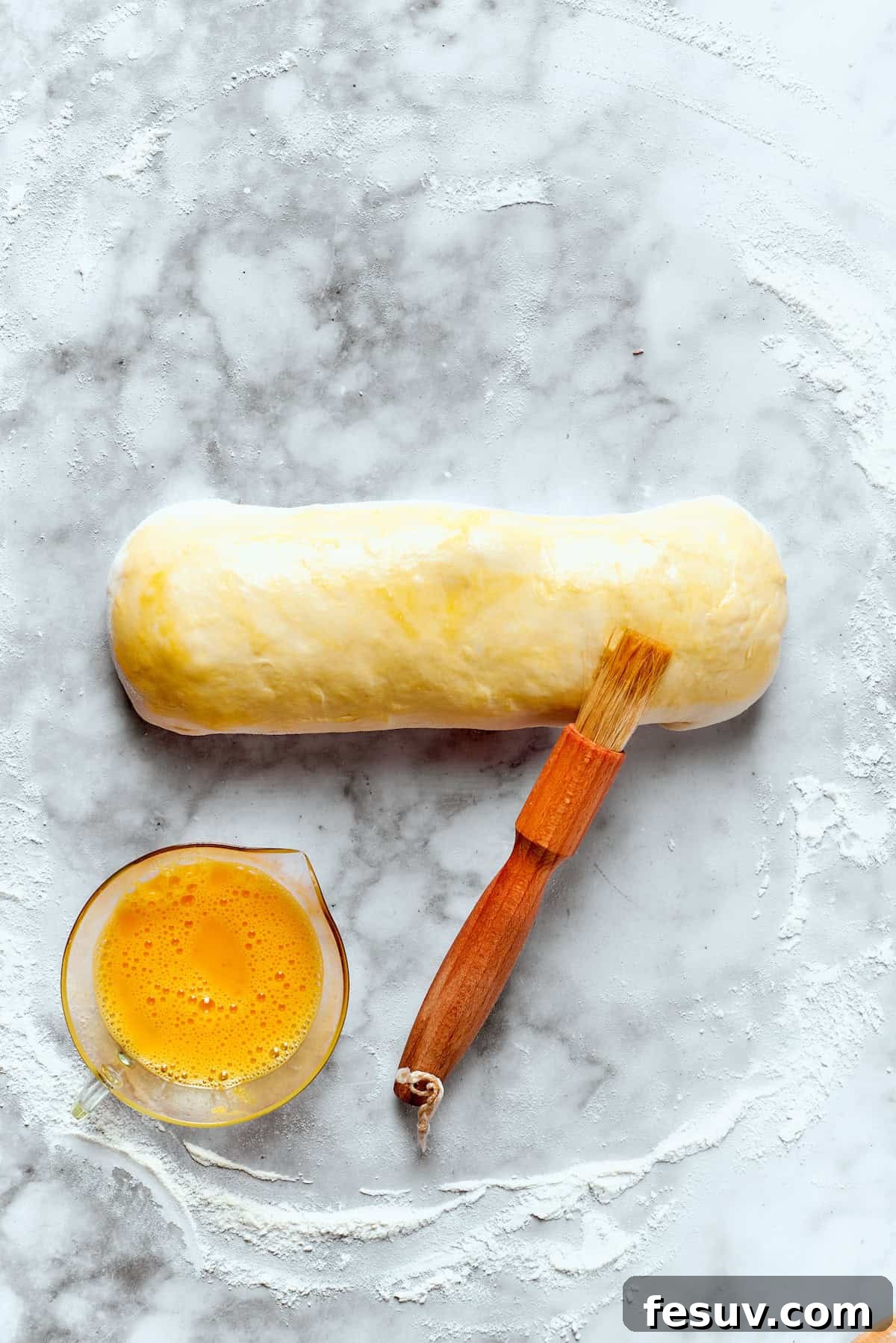 A rolled chicken bake pastry being brushed with egg wash on a baking sheet.