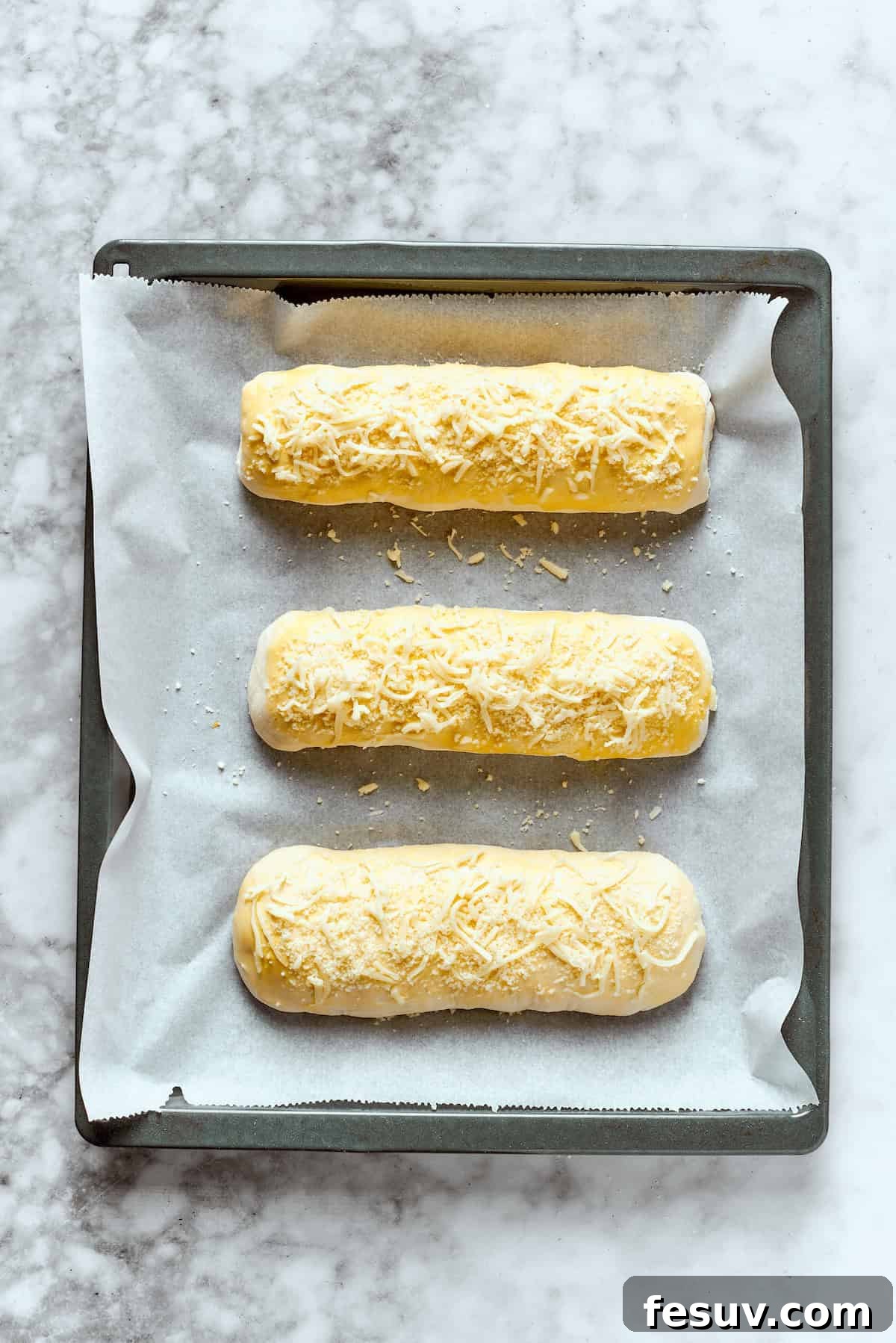 Three unbaked Costco chicken bake rolls arranged on a parchment-lined baking sheet, topped with cheese.