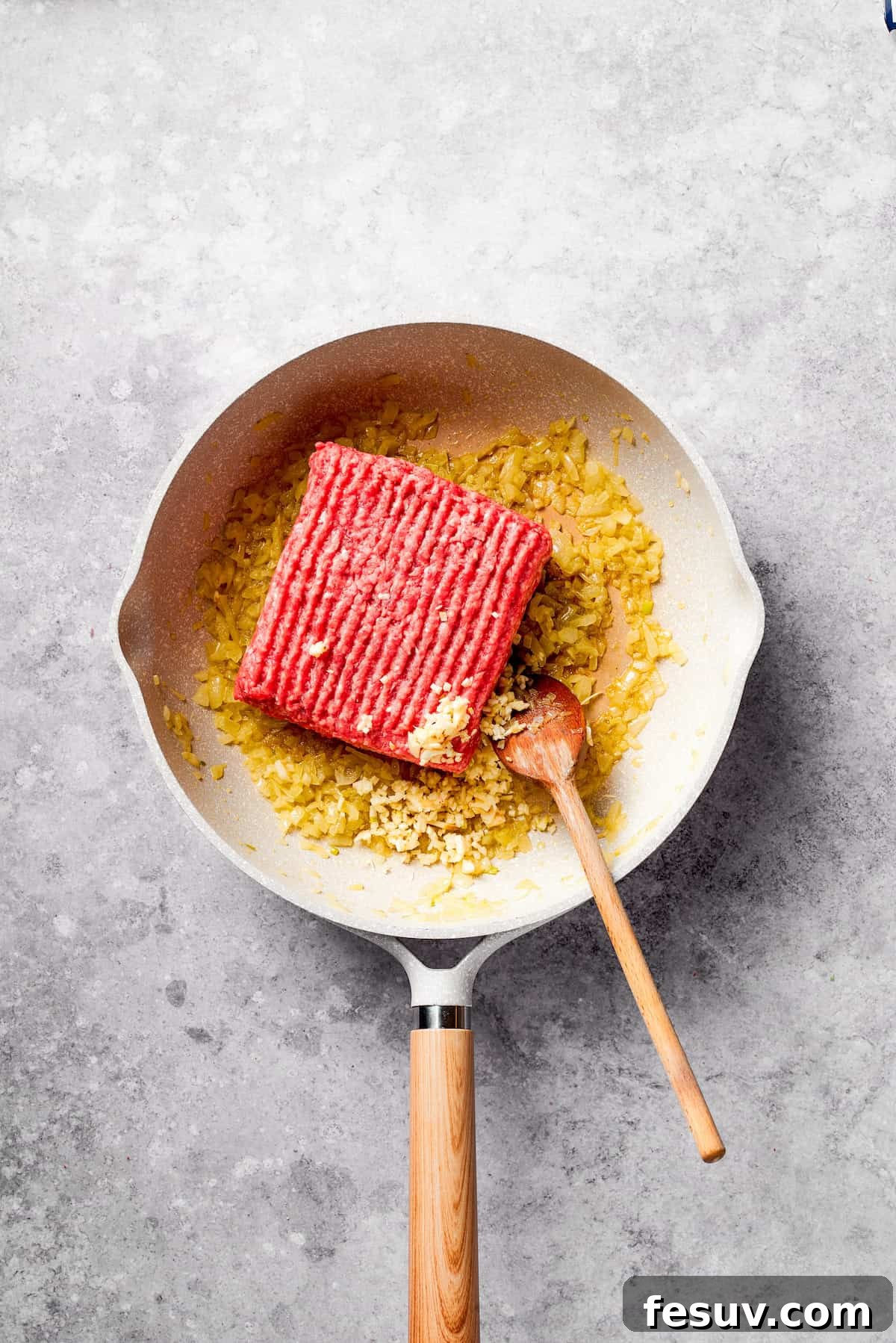The initial step of cooking diced onion and browning ground beef in a skillet. A block of ground beef being added to a hot frying pan with sautéed onions.