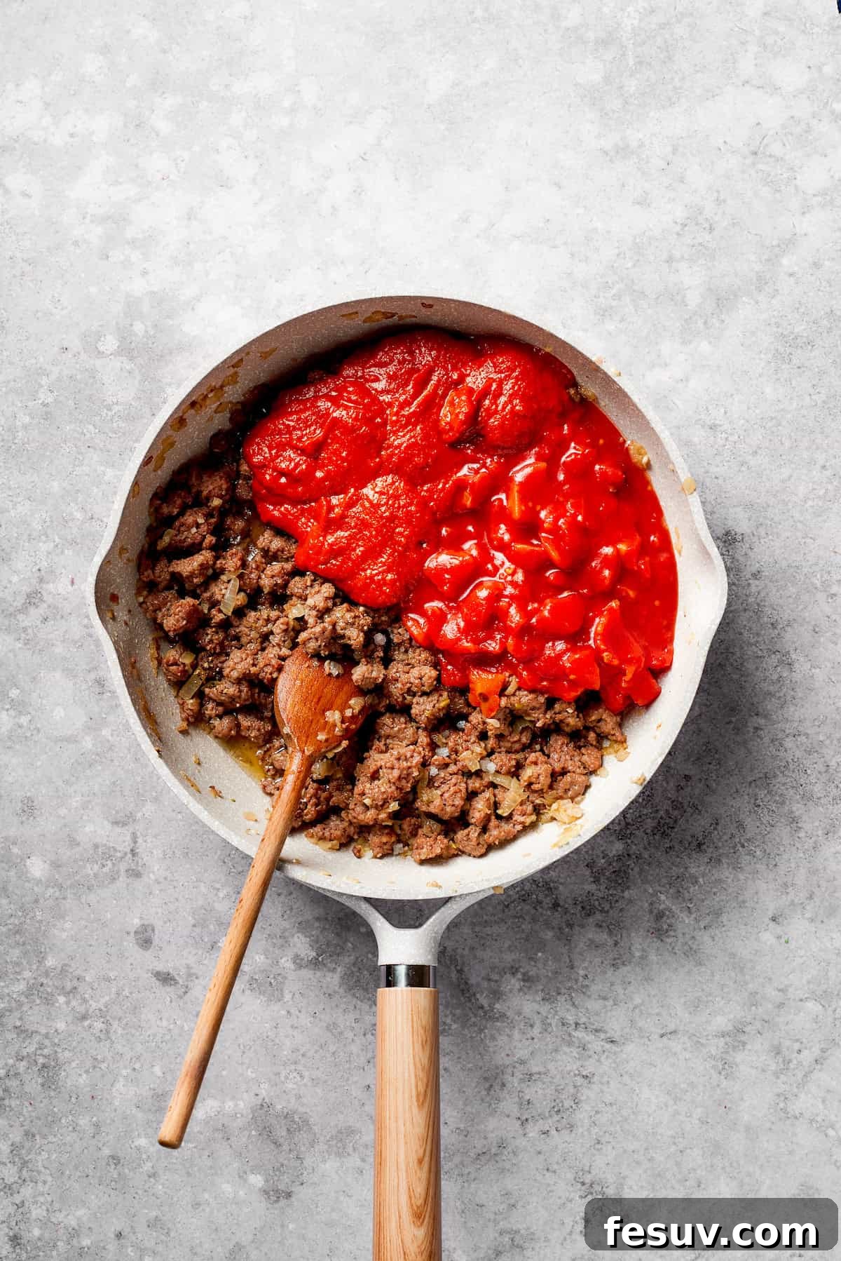 Adding canned tomatoes to the beef mixture to form the rich pasta sauce. Canned diced tomatoes being stirred into the browned ground beef mixture in a frying pan.