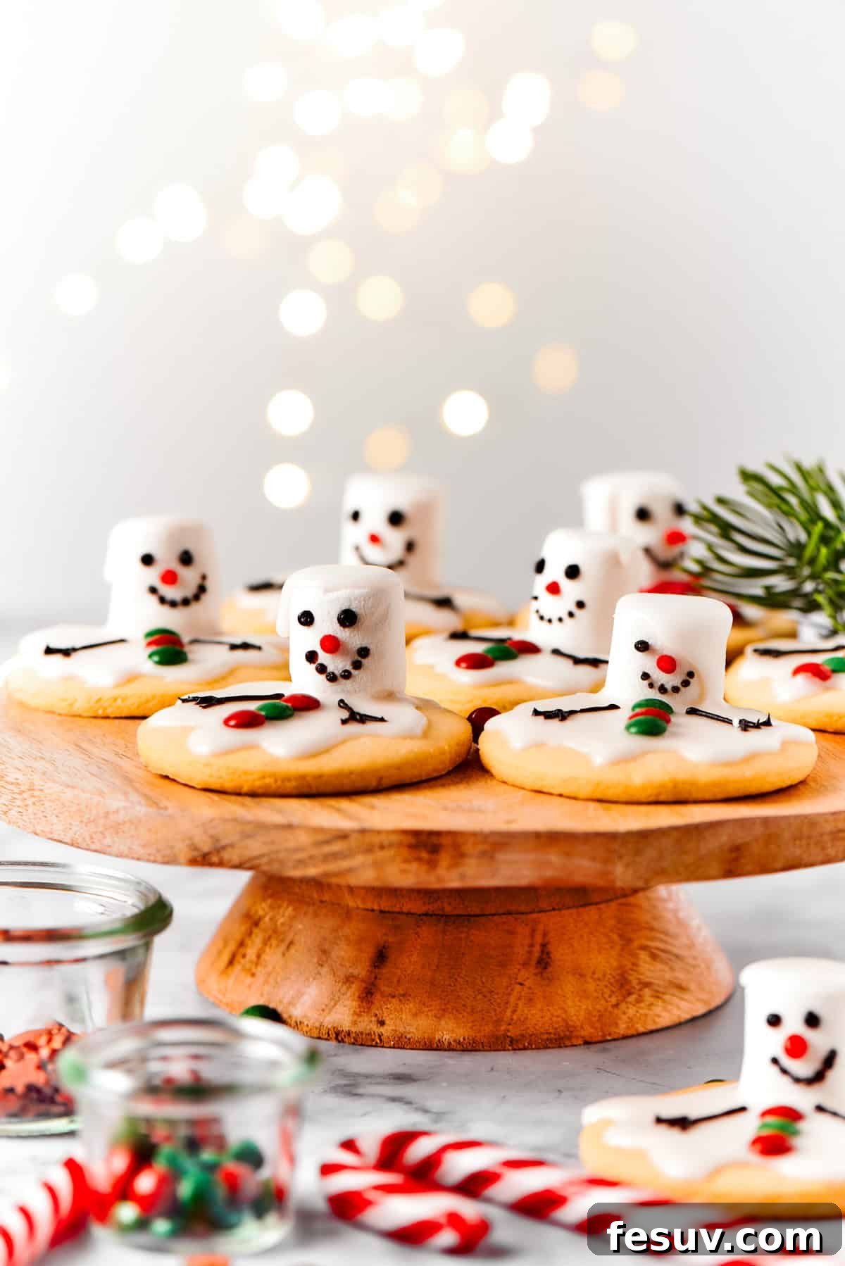 The cutest Melted Snowman Cookies top buttery homemade sugar cookies with a thick vanilla frosting and a fluffy marshmallow! Melting snowman cookies elegantly arranged on a rustic wooden cake plate, surrounded by charming holiday decor.