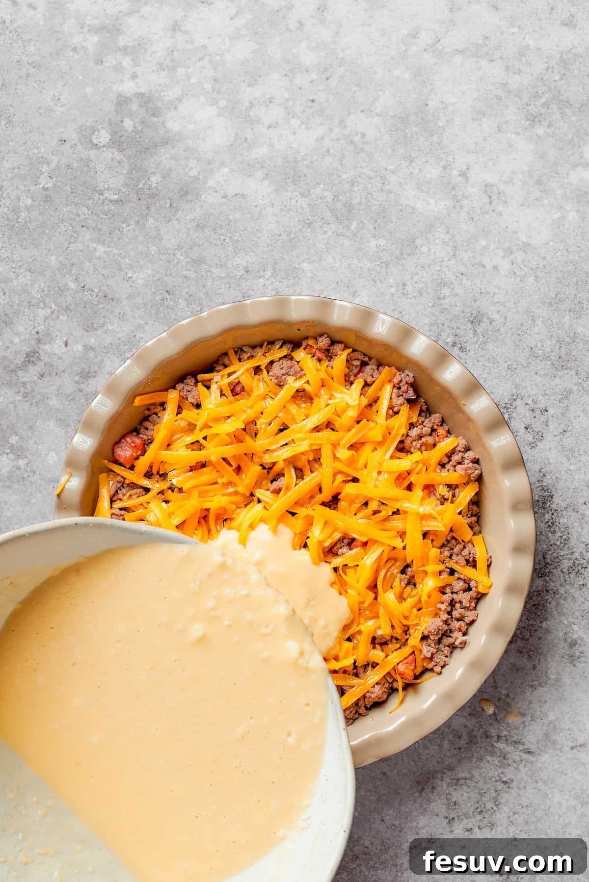 The prepared biscuit batter being carefully poured over the meat and cheese filling in the pie dish, creating the top crust.