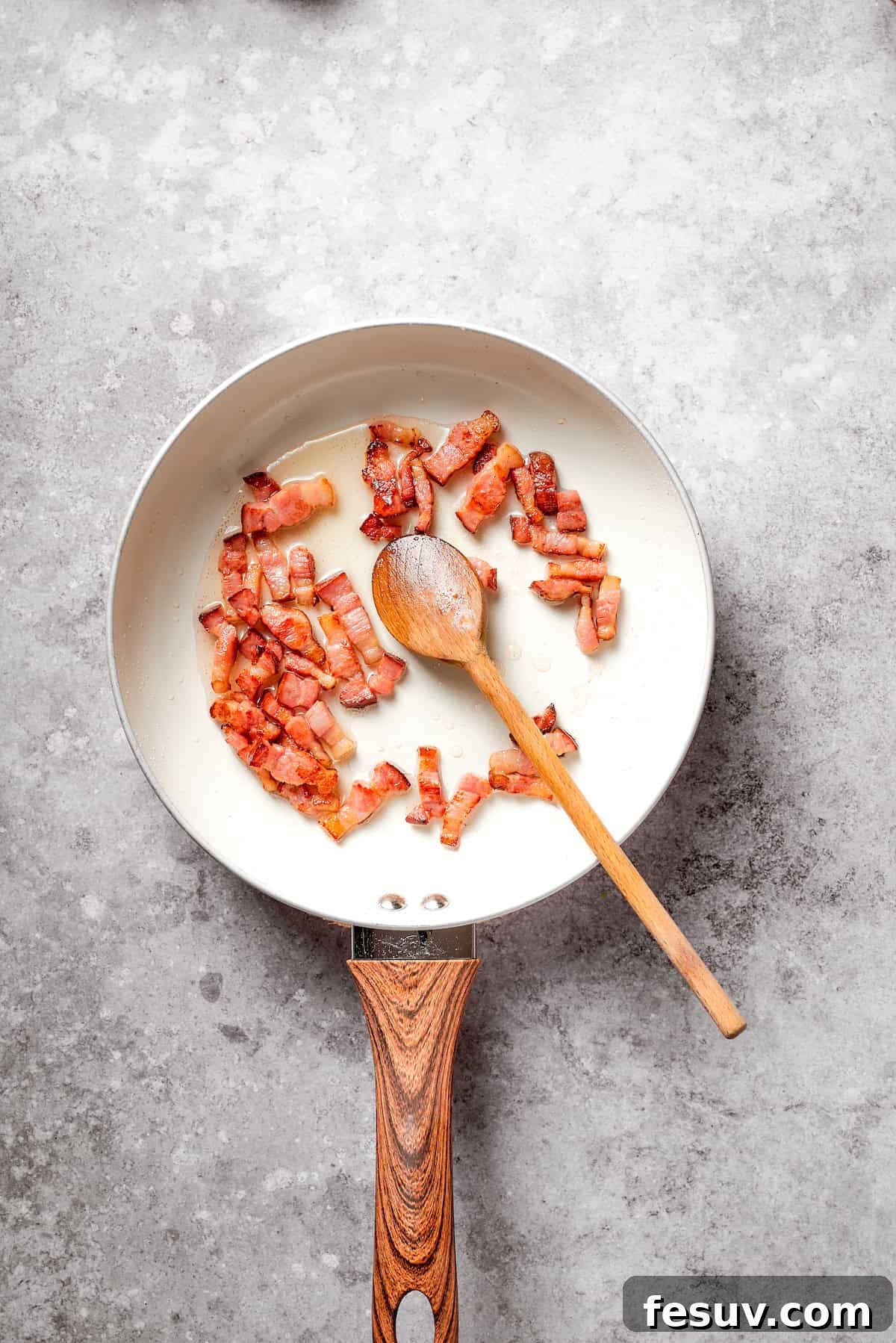 A spoon stirring chopped bacon as it cooks and crisps up in a skillet.