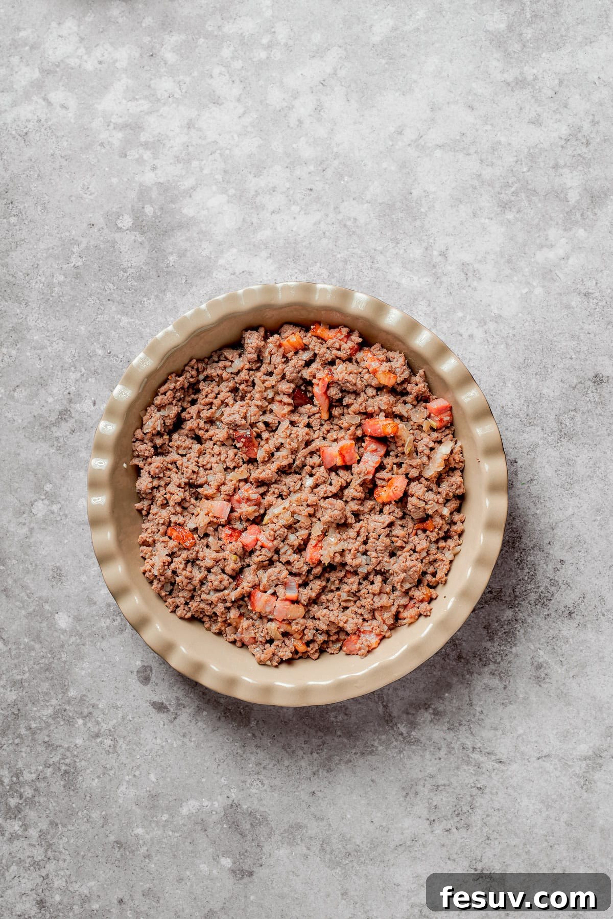 Prepared cheeseburger pie filling, consisting of seasoned ground beef and crispy bacon, neatly spread into a pie pan.
