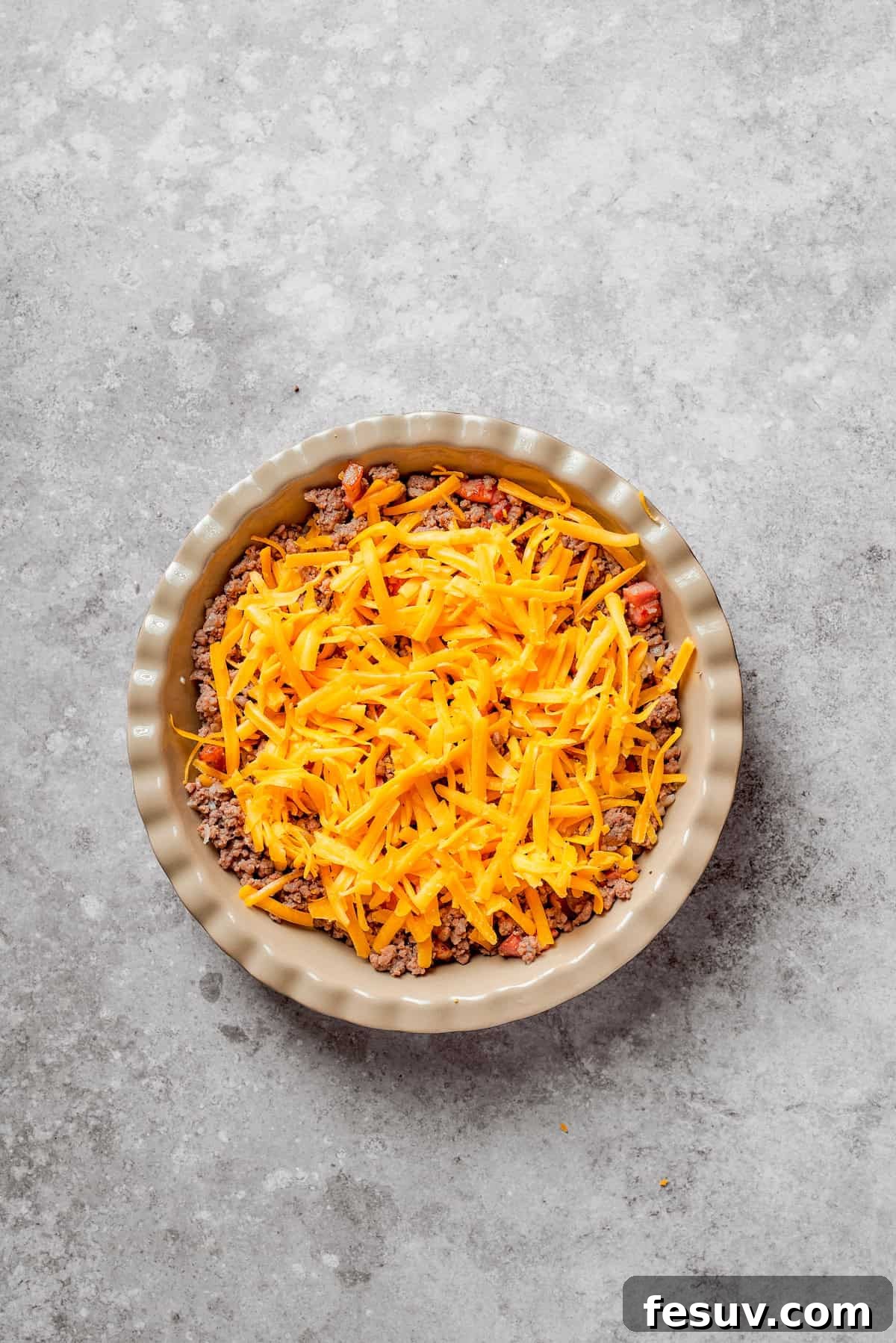 A generous layer of shredded cheddar cheese being sprinkled over the ground beef and bacon mixture in the pie pan.