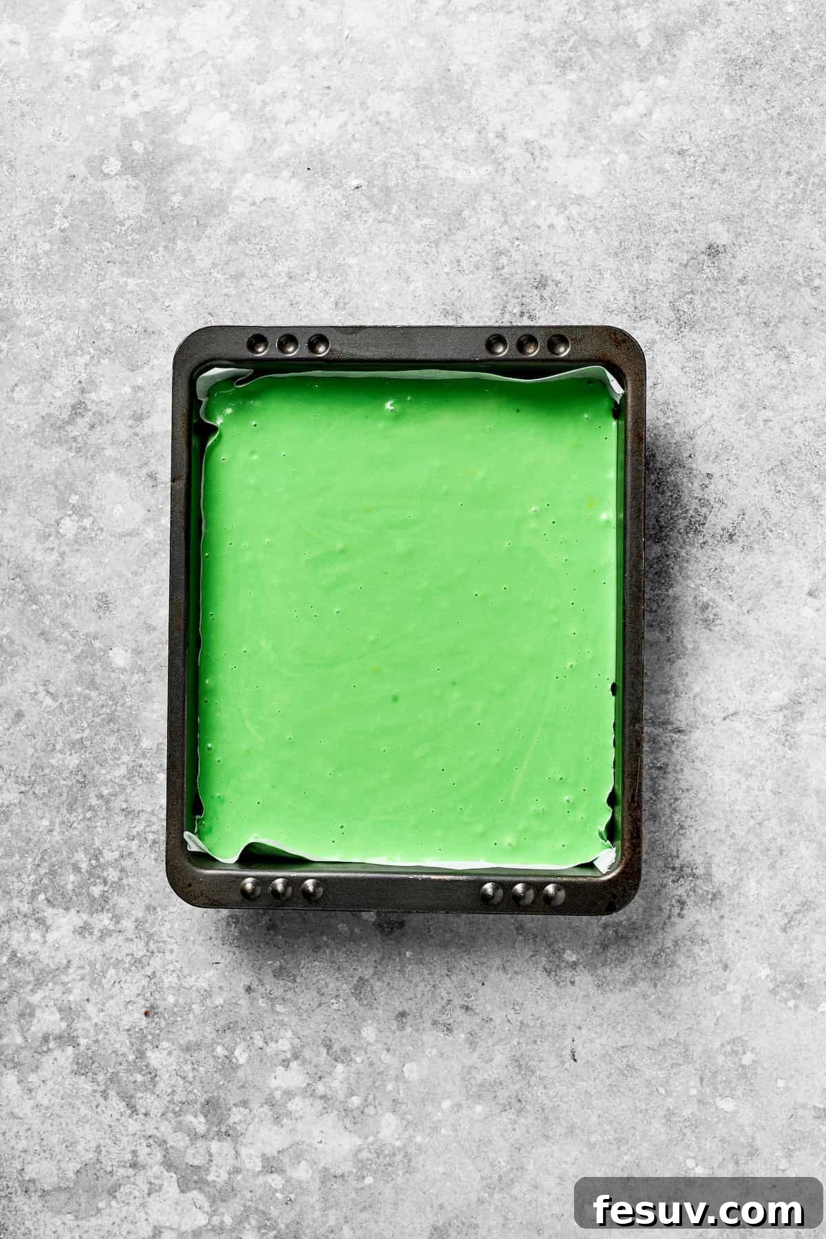 Green Grinch Cake features a tender, brightly-hued vanilla cake topped with tangy cream cheese frosting and studded with bits of candy. A square pan with green cake batter.