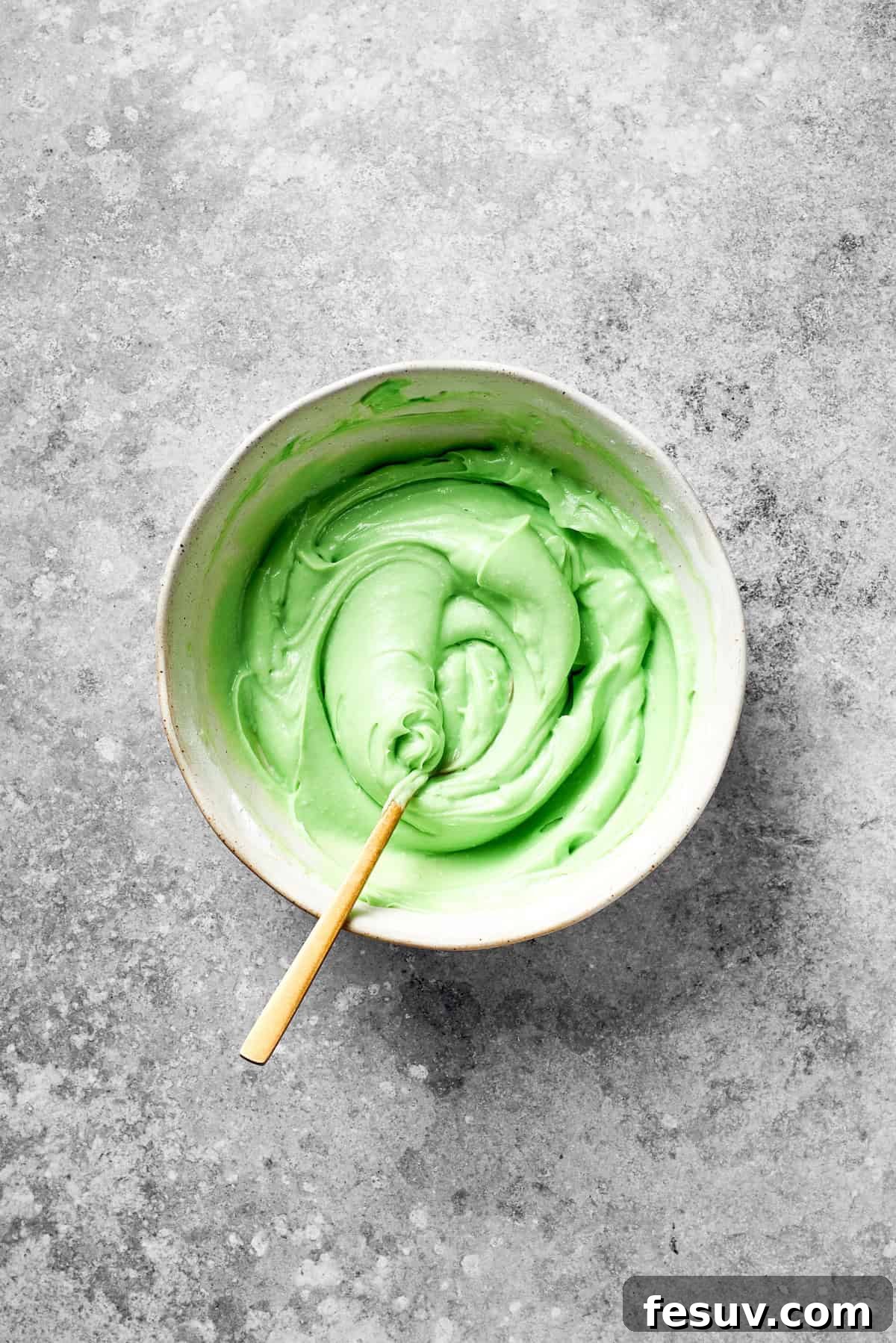 Green Grinch Cake features a tender, brightly-hued vanilla cake topped with tangy cream cheese frosting and studded with bits of candy. A spoon stirs green food coloring into icing.