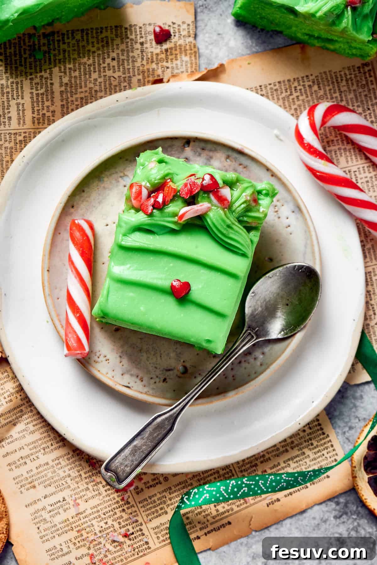 Green Grinch Cake features a tender, brightly-hued vanilla cake topped with tangy cream cheese frosting and studded with bits of candy. A plate with a slice of grinch cake and a spoon.