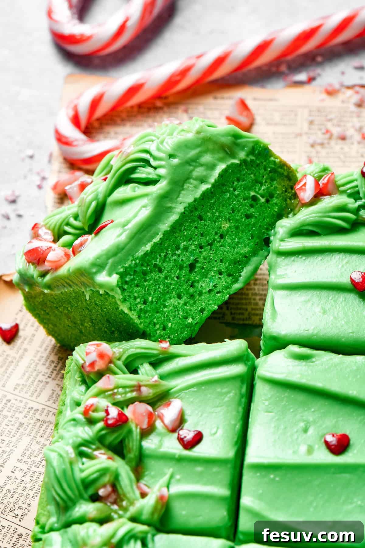 Green Grinch Cake features a tender, brightly-hued vanilla cake topped with tangy cream cheese frosting and studded with bits of candy. A slice of green cake turned on its side.