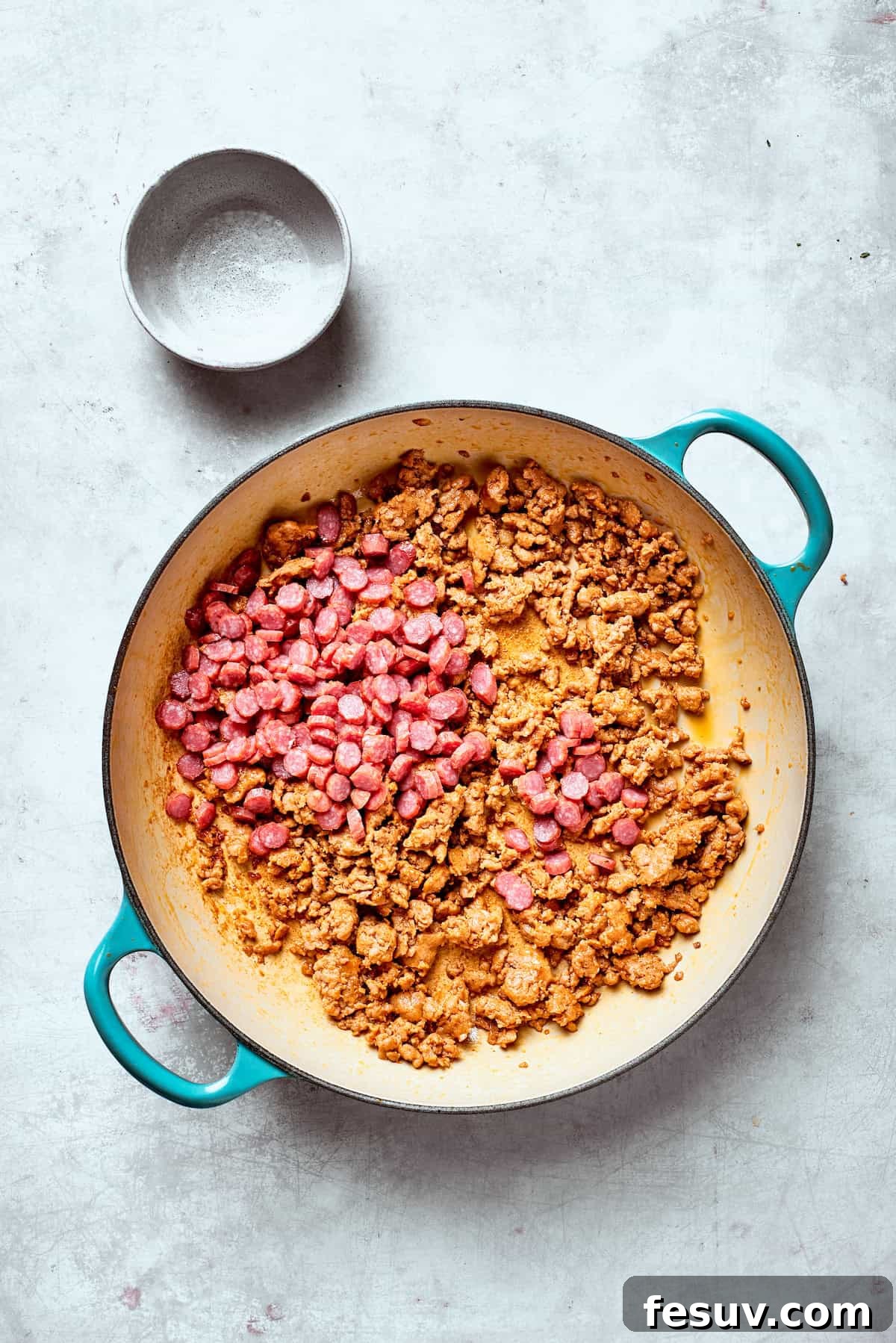 Pepperoni slices are added to a skillet containing browned and crumbled Italian sausage, ready for the next step.
